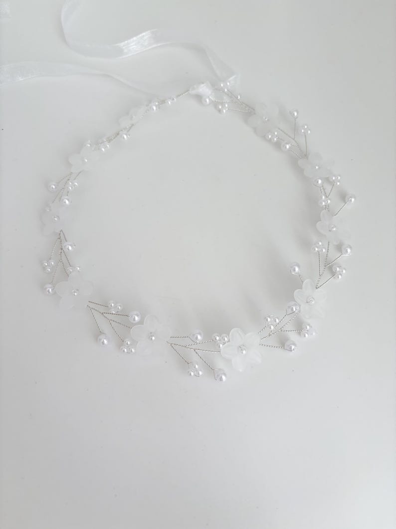 May include: A delicate white floral headband with faux pearls and a ribbon tie. The headpiece features small, translucent flowers and clusters of pearl beads, perfect for weddings or special occasions.