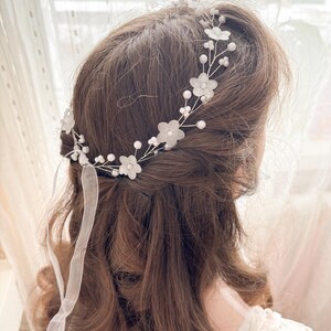 May include: A delicate floral hairpiece featuring white fabric flowers and small pearls. The headpiece is attached to a silver wire frame and includes sheer white ribbons. This bridal accessory is designed to be worn in the hair.
