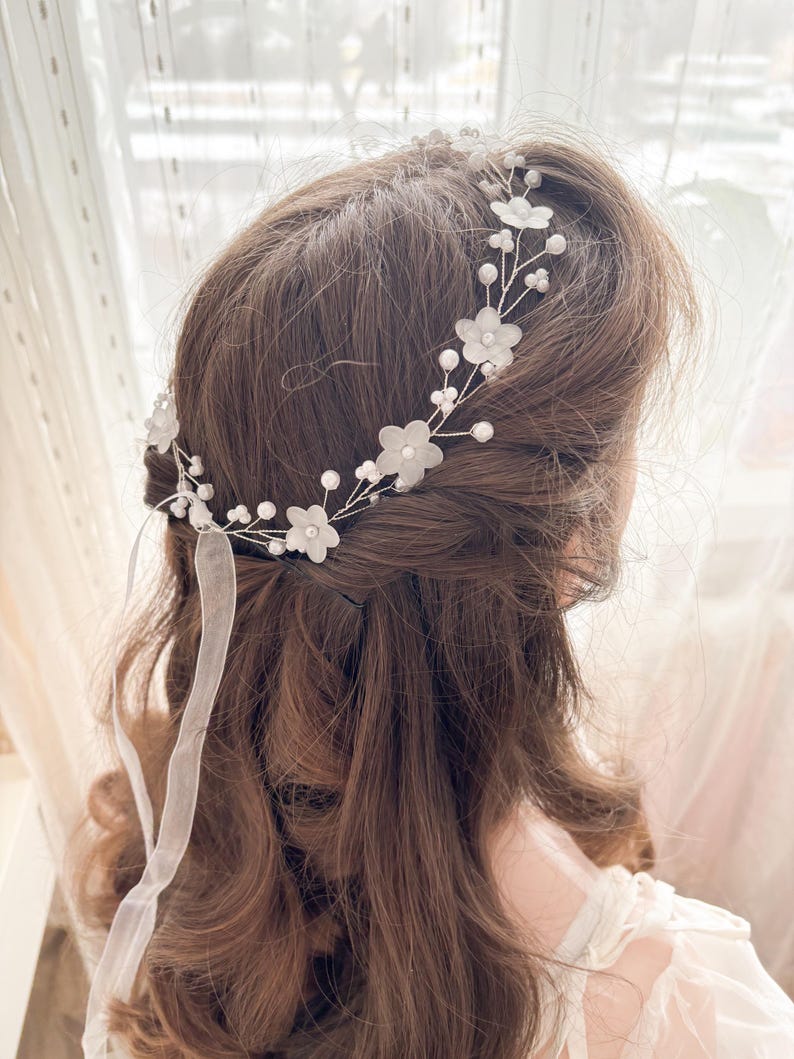 May include: A delicate floral hairpiece featuring white fabric flowers and small pearls. The headpiece is attached to a silver wire frame and includes sheer white ribbons. This bridal accessory is designed to be worn in the hair.