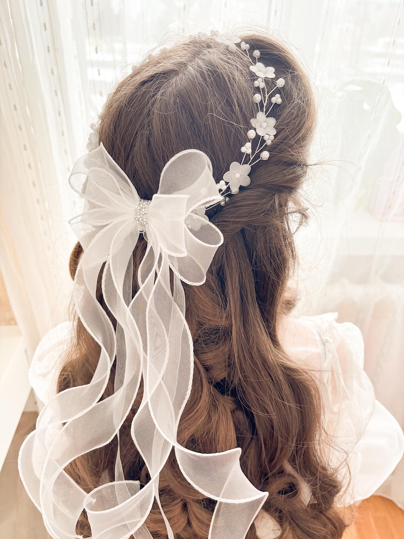 May include: A white bridal hair accessory featuring a large bow with long, flowing ribbons and a sparkling rhinestone center. Delicate floral and pearl accents adorn the hairpiece, creating an elegant and romantic look for weddings or special occasions.