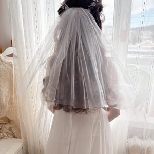 May include: A bride wearing a white wedding dress and veil, standing in front of a window. The veil is sheer and covers the back of the head and shoulders. A floral hairpiece is visible.