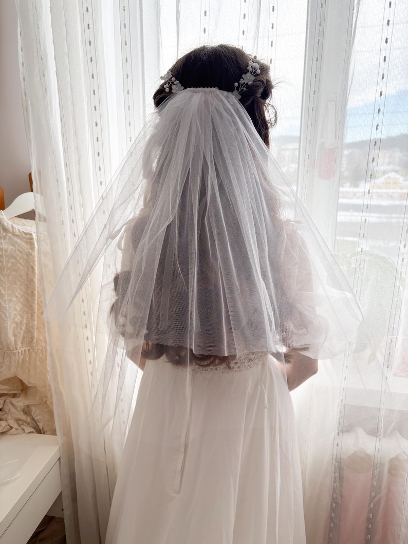 May include: A bride wearing a white wedding dress and veil, standing in front of a window. The veil is sheer and covers the back of the head and shoulders. A floral hairpiece is visible.