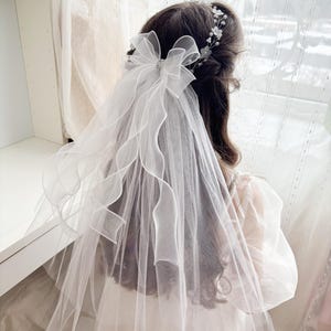May include: A white bridal veil with a large bow and a delicate floral headpiece. The veil is made of sheer fabric and drapes down the back. The bow has a sparkling embellishment in the center. The hair is dark brown.