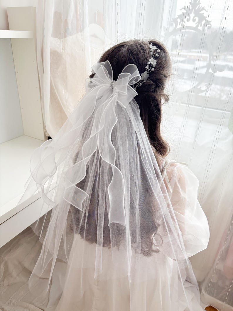 May include: A white bridal veil with a large bow and a delicate floral headpiece. The veil is made of sheer fabric and drapes down the back. The bow has a sparkling embellishment in the center. The hair is dark brown.