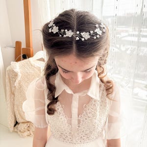 May include: A delicate floral hair crown featuring white fabric flowers and small pearl accents. The crown is set on a thin, silver-toned wire. The image shows a young person wearing the crown with a white dress.