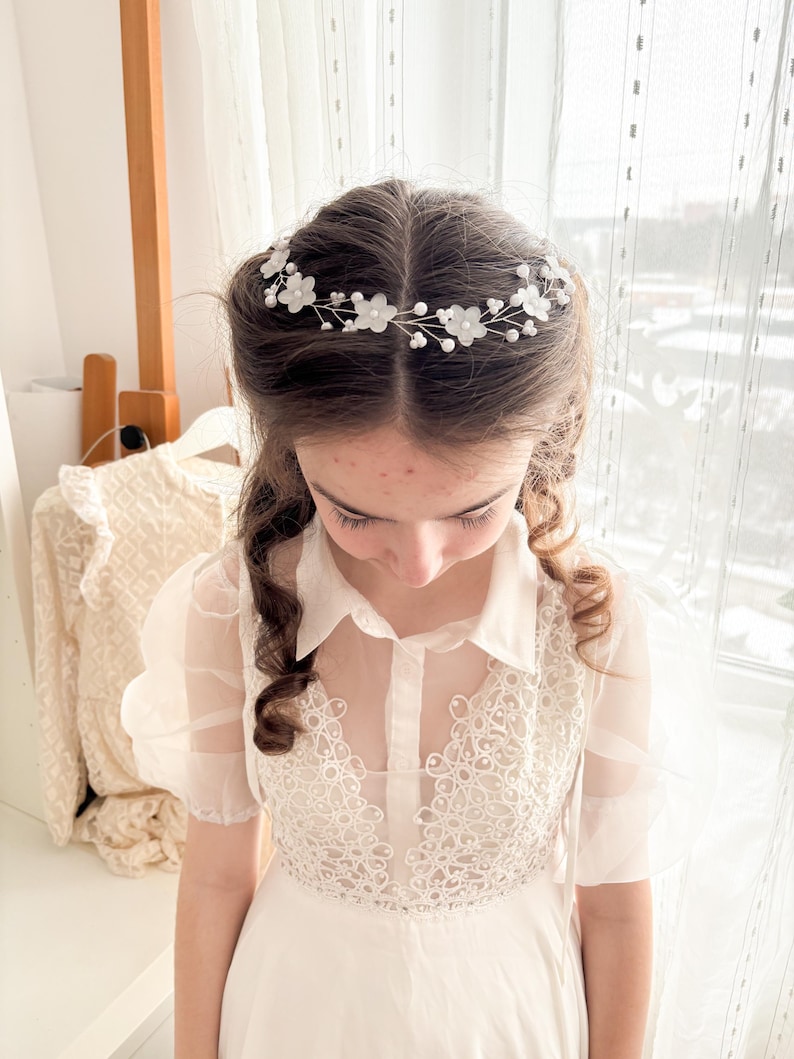 May include: A delicate floral hair crown featuring white fabric flowers and small pearl accents. The crown is set on a thin, silver-toned wire. The image shows a young person wearing the crown with a white dress.