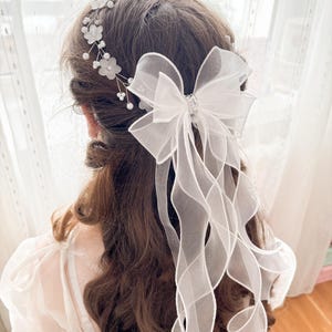 May include: A white bridal hair accessory featuring a large bow with a sparkling center and flowing ribbons. Delicate floral and pearl accents adorn the hairpiece, creating an elegant and romantic look. The accessory is designed to be worn in the hair.