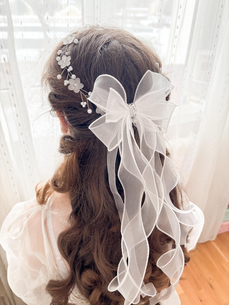May include: A white bridal hair accessory featuring a large bow with a sparkling center and flowing ribbons. Delicate floral and pearl accents adorn the hairpiece, creating an elegant and romantic look. The accessory is designed to be worn in the hair.