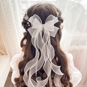 May include: A white organza hair bow with a sparkling rhinestone center is attached to the back of the hair. Long, flowing ribbons cascade down the back. The hair is styled in loose curls, with a delicate floral and pearl hairpiece.