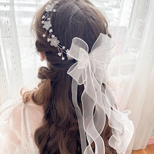 May include: A white bridal hair accessory featuring a floral headband and a large bow with flowing ribbons. The headband is adorned with small white flowers and pearls. The bow has a sparkling center.