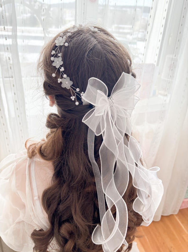 May include: A white bridal hair accessory featuring a floral headband and a large bow with flowing ribbons. The headband is adorned with small white flowers and pearls. The bow has a sparkling center.