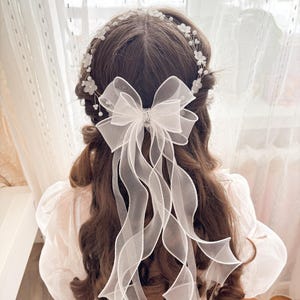 May include: A white bridal hair accessory featuring a delicate floral headband and a large sheer bow with long, flowing ribbons. The bow has a sparkling rhinestone center, adding a touch of elegance to the hairstyle. The accessory is designed for a wedding or special occasion.