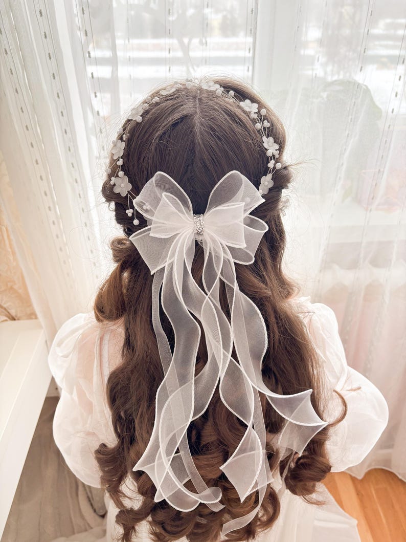 May include: A white bridal hair accessory featuring a delicate floral headband and a large sheer bow with long, flowing ribbons. The bow has a sparkling rhinestone center, adding a touch of elegance to the hairstyle. The accessory is designed for a wedding or special occasion.