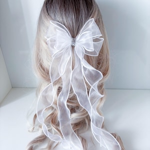 May include: A white organza bow hair accessory with a sparkling rhinestone center. The bow features long, flowing ribbons that cascade down. The hair accessory is set against a backdrop of blonde and brown hair.