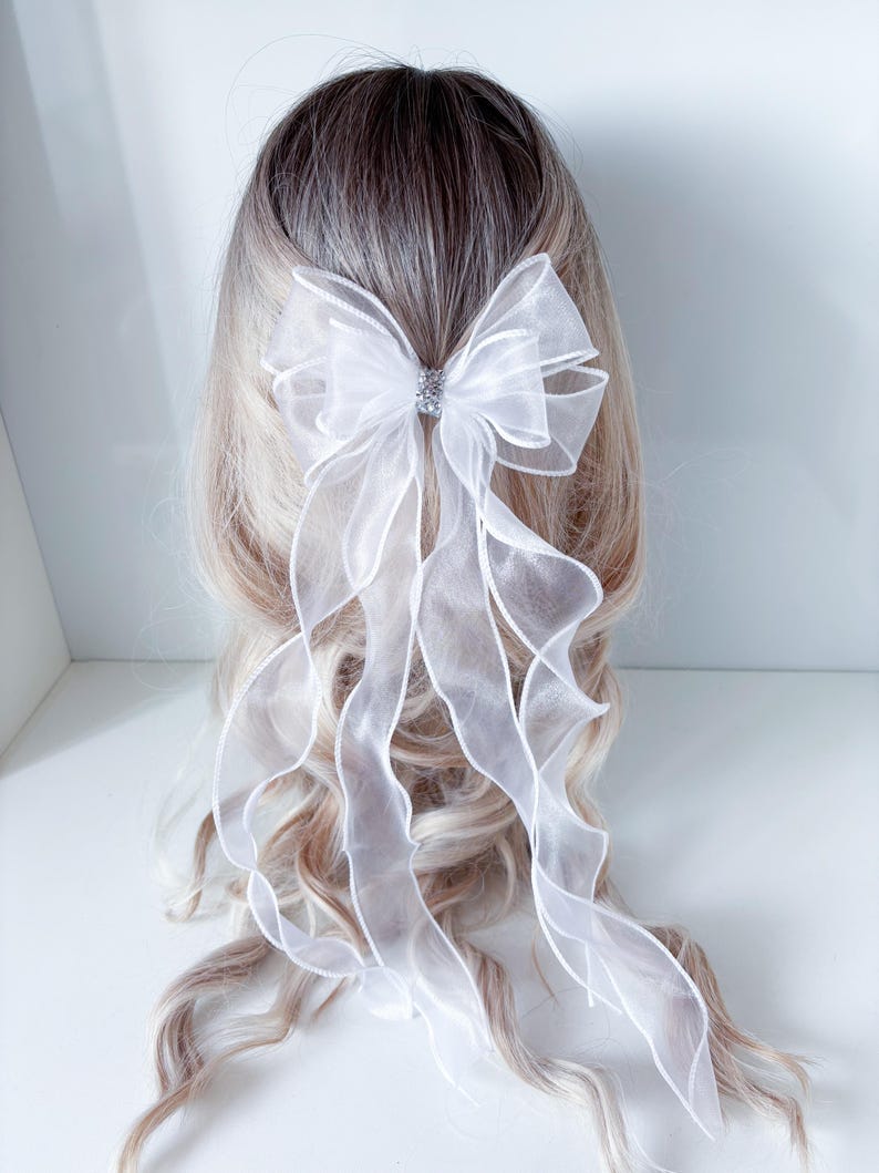 May include: A white organza bow hair accessory with a sparkling rhinestone center. The bow features long, flowing ribbons that cascade down. The hair accessory is set against a backdrop of blonde and brown hair.