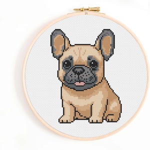 Pode incluir: Bordado em ponto cruz de um filhote de buldogue francês em um bastidor de madeira. O cão é bege com detalhes em preto e cinza, e sua língua é rosa. O fundo é branco.