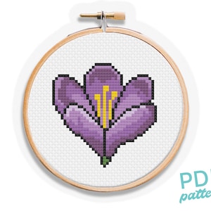 May include: A cross-stitch pattern of a purple flower with yellow stamens, displayed in a wooden embroidery hoop. The flower is pixelated, with a green stem. The text "PDF pattern" is in the lower right corner.