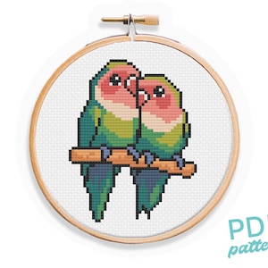 Può includere: Schema di punto croce di due tortore appollaiate su un ramo, con colori verde, rosa e bianco. Lo schema è in un telaio da ricamo in legno.