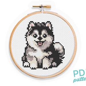 May include: Cross stitch pattern of a cute, cartoon husky puppy with black and white fur, sitting with its tongue sticking out. The pattern is in a wooden embroidery hoop.