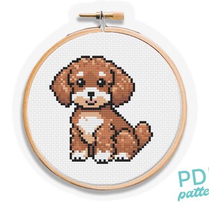 Puede incluir: Patrón de punto de cruz de un cachorro marrón y blanco sentado sobre un fondo blanco. El patrón está en un aro de bordado de madera redondo.