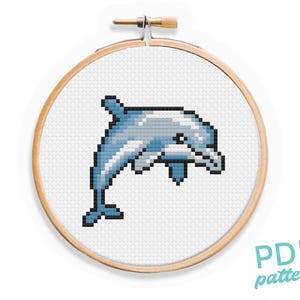 May include: A cross-stitch pattern featuring a blue and grey dolphin jumping out of the water. The pattern is displayed in a wooden embroidery hoop.