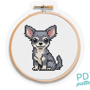 以下が含まれることがあります： 白い背景にピンクの耳をしたグレーとホワイトのチワワが座っているクロスステッチパターン。パターンは木製の刺繍枠に入っています。