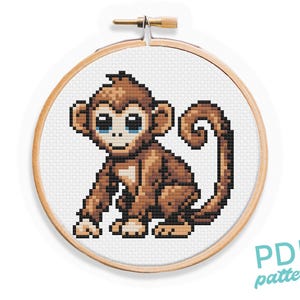 May include: Cross stitch pattern of a brown monkey with blue eyes. The monkey is sitting with its tail curled around its body. The pattern is in a wooden embroidery hoop.