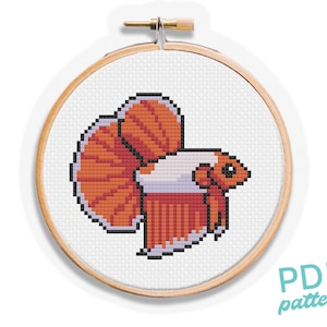 May include: A cross-stitch pattern of a betta fish in an embroidery hoop. The fish features orange and white pixelated details, with a prominent tail and fins. The text "PDF pattern" is visible in the lower right corner.