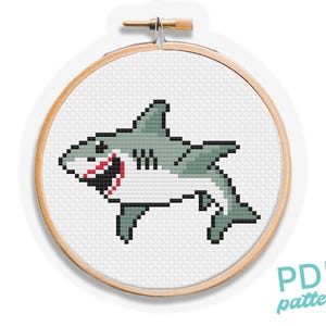 May include: A cross stitch pattern featuring a pixel art shark design in shades of gray, white, and black with a red mouth, displayed within a circular wooden embroidery hoop. The design is presented as a digital PDF pattern for needlework.