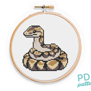 May include: A cross-stitch pattern featuring a coiled snake with brown, tan, and white scales. The snake is curled up in a circle, with its head facing the viewer. The pattern is in a wooden embroidery hoop.
