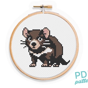 May include: Cross stitch pattern of a brown and black Tasmanian devil with pink ears. The pattern is in a round wooden embroidery hoop.