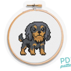 Puede incluir: Un patrón de punto de cruz de un perro Cavalier King Charles Spaniel en un bastidor de bordado de madera. El perro está representado en estilo pixel art, con colores negro, marrón y beige. El texto "PDF pattern" está en la esquina inferior derecha.