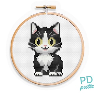 Peut inclure: Un chat noir et blanc aux yeux jaunes est assis dans un cercle de broderie rond. Le chat a une tache blanche sur la poitrine et un nez rose.