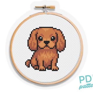Puede incluir: Un patrón de punto de cruz de un perro Cavalier King Charles Spaniel en un bastidor de bordado de madera. El perro está representado en tonos marrones y negros, con un diseño pixelado. El texto "PDF pattern" está en la esquina inferior derecha.