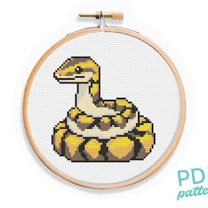 Peut inclure: Un motif de point de croix représentant un serpent jaune et brun enroulé en boule. Le motif est affiché sur un tambour à broder en bois.