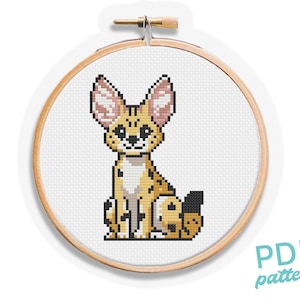May include: Cross stitch pattern of a cute, cartoon-style, yellow and brown,  caracal with pink ears sitting on a white background. The pattern is in a round wooden embroidery hoop.