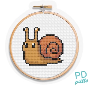 May include: A cross-stitch pattern of a pixelated snail in shades of brown and orange, set within a wooden embroidery hoop. The design is on white fabric. The text "PDF pattern" is visible.