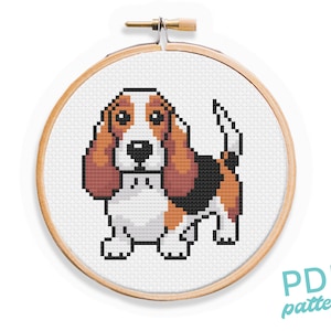 May include: Cross stitch pattern of a brown and white basset hound dog with floppy ears. The pattern is displayed in a wooden embroidery hoop.