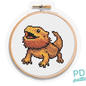 May include: A cross-stitch pattern of a bearded dragon in a wooden embroidery hoop. The lizard is rendered in shades of orange, brown, and black, with its mouth open. The text "PDF pattern" is in the lower right corner.