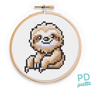 May include: A cross-stitch pattern featuring a cute brown and white sloth with a happy expression. The pattern is displayed in a wooden embroidery hoop.