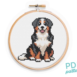 May include: Cross stitch pattern of a Bernese Mountain Dog sitting with its tongue sticking out. The dog is black, white, and brown. The pattern is in a wooden embroidery hoop.
