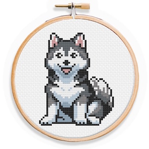 May include: Cross-stitch embroidery of a black and white husky dog with pink ears and a light blue tongue. The dog is sitting with its front paws crossed and its tail curled behind it. The embroidery is framed in a round wooden hoop.