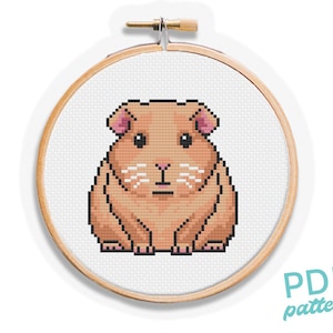 May include: Cross stitch pattern of a brown and white guinea pig. The guinea pig is sitting with its paws tucked under its body. The pattern is in a white embroidery hoop.