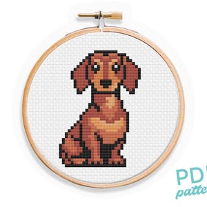 May include: Cross stitch pattern of a brown dachshund dog sitting with its ears perked up. The pattern is in a round wooden embroidery hoop.