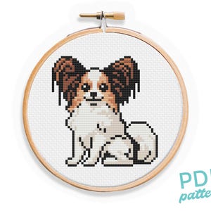 May include: Cross stitch pattern of a Papillon dog sitting with its ears perked up. The dog is white with brown ears and a brown patch on its back. The pattern is in a wooden embroidery hoop.