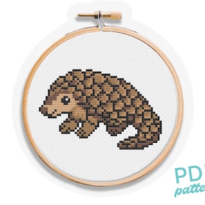 Può includere: Un modello a punto croce raffigurante un pangolino in pixel art nei toni del marrone e del beige su tessuto bianco, fissato all'interno di un telaio da ricamo circolare in legno. Il disegno mostra un animale stilizzato con corpo squamato.