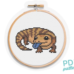 Könnte beinhalten: Ein Kreuzstichmuster eines Echsen in einem hölzernen Stickrahmen. Die Echse ist braun und beige mit einer blauen Zunge. Der Text "PDF pattern" befindet sich in der unteren rechten Ecke.