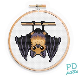May include: Cross stitch pattern of a brown bat with purple wings hanging upside down from a branch. The pattern is in a white embroidery hoop.