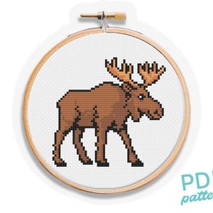 May include: A cross-stitch pattern of a moose in shades of brown and tan, displayed in a wooden embroidery hoop. The moose is pixelated, giving it a retro look. The text "PDF pattern" is in the lower right corner.