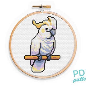 May include: A cross-stitch design featuring a white cockatoo bird on a brown branch, framed in a wooden embroidery hoop. The bird has yellow crest feathers and is created in a pixelated style. The text "PDF pattern" is visible in the lower right corner.
